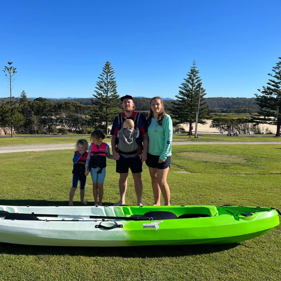 dalmeny campground family standing in grassed area with a paddle board and beach in the background